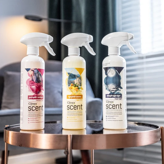 Trzy butelki odświeżaczy powietrza Clinex Scent o zapachach Paryski wdzięk, Egejski owoc oraz Magiczna noc, stojące na szklanym stole.