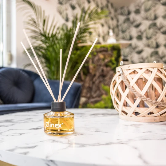 Buteleczka z patyczkami zapachowymi Clinex Scent Spirit stoi na marmurowym stoliku obok drewnianego świecznika. W tle widoczna jest granatowa sofa i kwiat, co tworzy elegancką aranżację wnętrza.