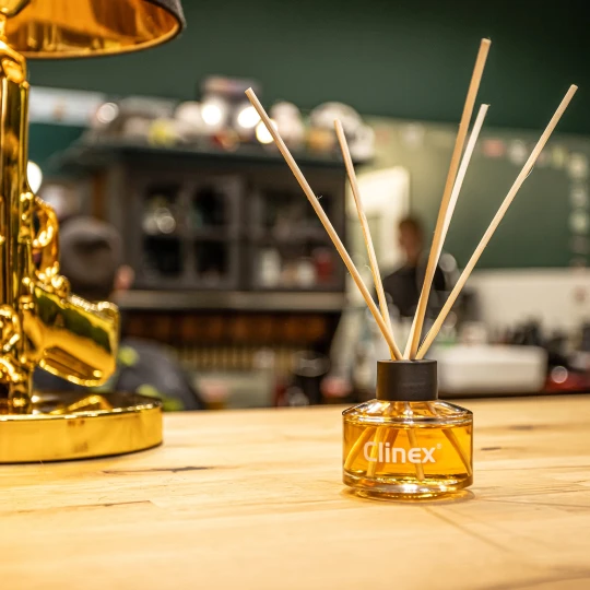 Buteleczka z patyczkami zapachowymi Clinex Scent Spirit stoi na drewnianym blacie w salonie fryzjerskim. W tle widoczne są ciemnozielone ściany, co podkreśla, że produkt jest przeznaczony do profesjonalnych i stylowych wnętrz.