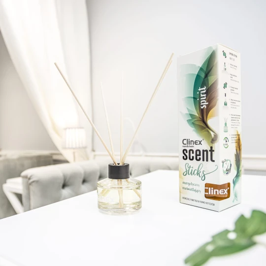 Buteleczka z patyczkami zapachowymi Clinex Scent Spirit stoi na białym blacie w salonie kosmetycznym. W tle widoczne są białe zasłony i szare fotele, co podkreśla, że produkt jest przeznaczony do profesjonalnych wnętrz.