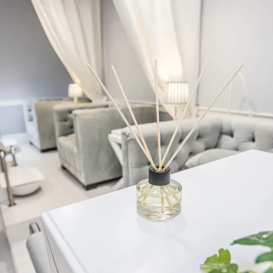 Buteleczka z patyczkami zapachowymi Clinex Scent Spirit stoi na białym blacie w salonie kosmetycznym. W tle widoczne są białe zasłony i szare fotele, co podkreśla, że produkt jest przeznaczony do profesjonalnych wnętrz.