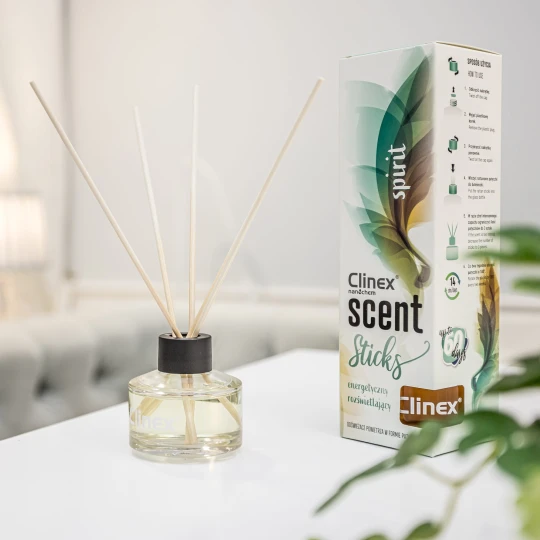 Buteleczka z patyczkami zapachowymi Clinex Scent Spirit stoi na białym blacie w salonie kosmetycznym. W tle widoczne są białe zasłony i szare fotele, co podkreśla, że produkt jest przeznaczony do profesjonalnych wnętrz.