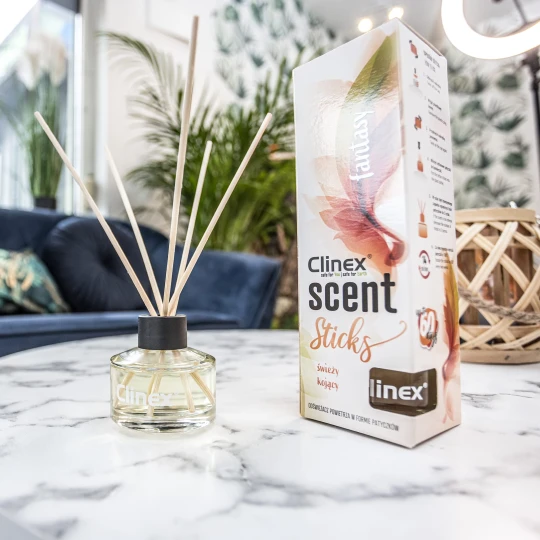 Buteleczka z patyczkami zapachowymi Clinex Scent Fastnasy stoi na marmurowym stoliku obok drewnianego świecznika. W tle widoczna jest granatowa sofa i kwiat, co tworzy elegancką aranżację wnętrza.