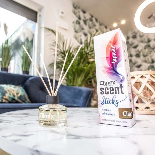 Buteleczka z patyczkami zapachowymi Clinex Scent Hipnotic stoi na marmurowym stoliku obok drewnianego świecznika. W tle widoczna jest granatowa sofa i kwiat, co tworzy elegancką aranżację wnętrza.