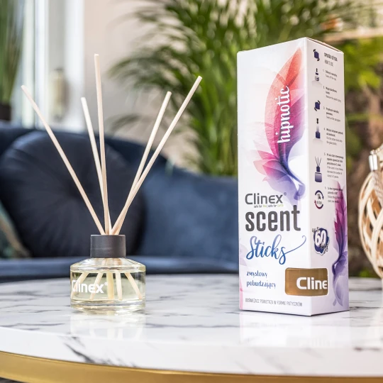 Buteleczka z patyczkami zapachowymi Clinex Scent Hipnotic stoi na marmurowym stoliku obok drewnianego świecznika. W tle widoczna jest granatowa sofa i kwiat, co tworzy elegancką aranżację wnętrza.