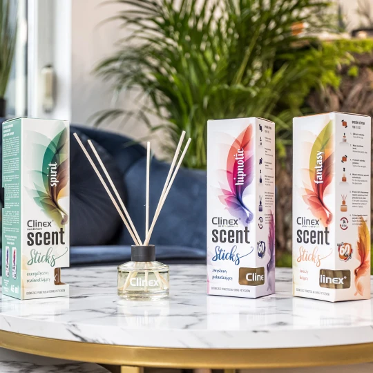 Trzy buteleczki z patyczkami zapachowymi Clinex Scent, w wariantach Spirit, Fastnasy i Hipnotic, stoją na marmurowym stoliku. W tle widoczna jest granatowa sofa i kwiat, co podkreśla, że produkt pasuje do eleganckiego wystroju wnętrz.