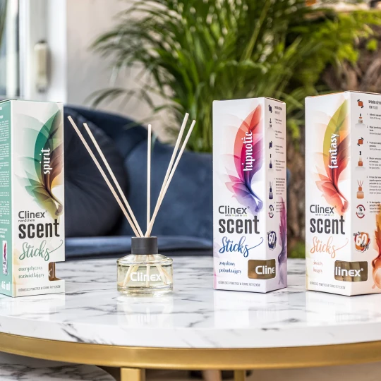 Trzy buteleczki z patyczkami zapachowymi Clinex Scent, w wariantach Spirit, Fastnasy i Hipnotic, stoją na marmurowym stoliku. W tle widoczna jest granatowa sofa i kwiat, co podkreśla, że produkt pasuje do eleganckiego wystroju wnętrz.