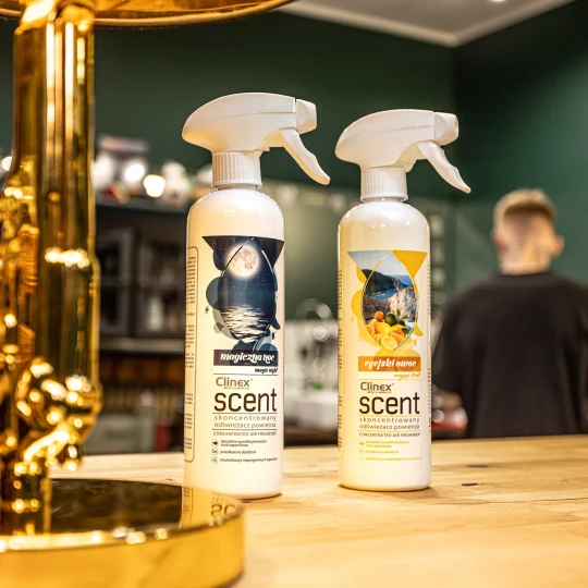 Dwie butelki odświeżacza powietrza Clinex Scent o zapachach Magiczna Noc i Egejski Owoc stoją na drewnianym blacie w salonie fryzjerskim, co podkreśla, że produkt jest przeznaczony do profesjonalnych wnętrz.