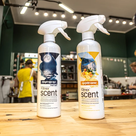 Dwie butelki odświeżacza powietrza Clinex Scent o zapachach Magiczna Noc i Egejski Owoc stoją na drewnianym blacie w salonie fryzjerskim, co podkreśla, że produkt jest przeznaczony do profesjonalnych wnętrz.
