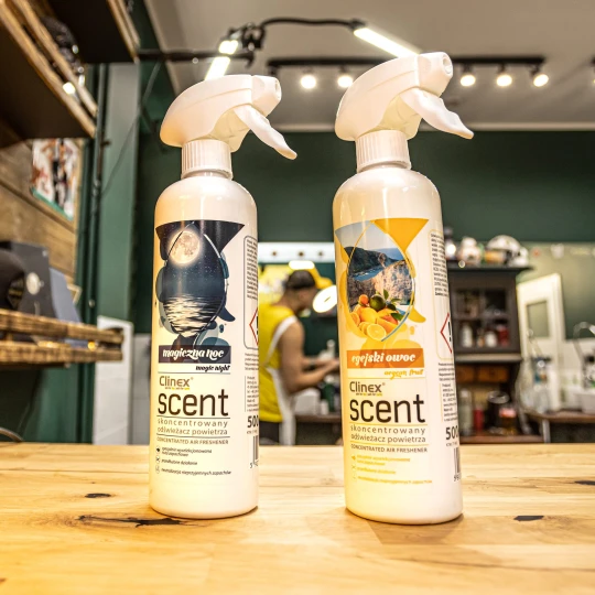 Dwie butelki odświeżacza powietrza Clinex Scent o zapachach Magiczna Noc i Egejski Owoc stoją na drewnianym blacie w salonie fryzjerskim, co podkreśla, że produkt jest przeznaczony do profesjonalnych wnętrz.