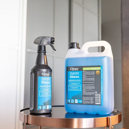Butelka Clinex Nano Protect Glass 1l oraz większy baniak o pojemności 5 litrów stoją na szklanym stoliku przed lustrem, co ukazuje dostępność produktu do mycia i zabezpieczania szkła w różnych formatach dla użytku domowego i profesjonalnego.