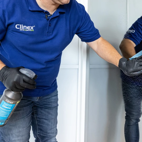 Osoba trzyma w ręce butelkę profesjonalnego płynu do mycia i zabezpieczania szkła Clinex Nano Protect Glass 1l i wyciera lustro niebieską ściereczką, co ukazuje sposób użycia produktu.