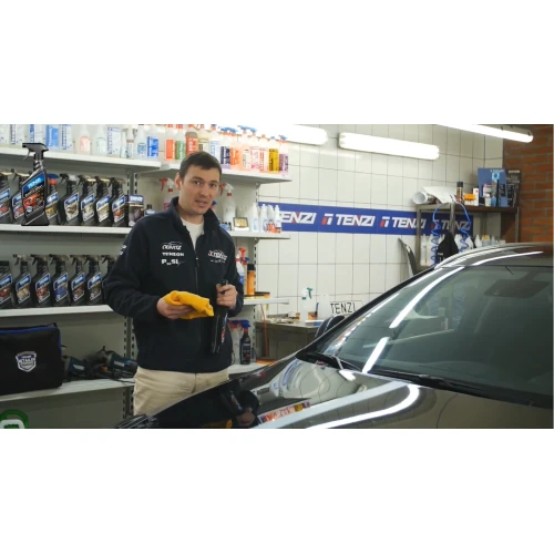 Mężczyzna stoi przed samochodem, trzymając w ręku butelkę quick detailera kwarcowego TENZI Detailer Quartz Spray oraz miękką ściereczkę, co symbolizuje przygotowanie do szybkiego nabłyszczania i zabezpieczania lakieru.