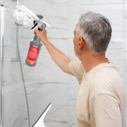 Mężczyzna spryskuje szklaną ścianę kabiny prysznicowej butelką preparatu Clinex W3 Active Shield, który skutecznie usuwa osady i chroni powierzchnię.