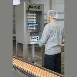 Realistyczna scena w nowoczesnym zakładzie przetwórstwa: pracownik obsługuje myjkę BATCH WASHER Mach-C, na obudowie widoczna etykieta produktu, w tle linia produkcyjna z transportowanymi jajkami oraz elementy hali podkreślające profesjonalne zastosow