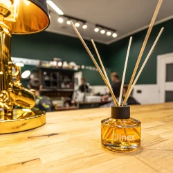 Buteleczka z patyczkami zapachowymi Clinex Scent Spirit stoi na drewnianym blacie w salonie fryzjerskim. W tle widoczne są ciemnozielone ściany, co podkreśla, że produkt jest przeznaczony do profesjonalnych i stylowych wnętrz.