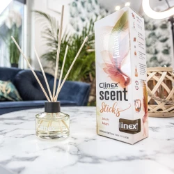 Buteleczka z patyczkami zapachowymi Clinex Scent Fastnasy stoi na marmurowym stoliku obok drewnianego świecznika. W tle widoczna jest granatowa sofa i kwiat, co tworzy elegancką aranżację wnętrza.