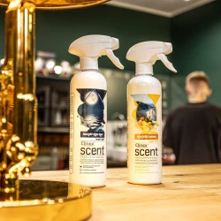 Dwie butelki odświeżacza powietrza Clinex Scent o zapachach Magiczna Noc i Egejski Owoc stoją na drewnianym blacie w salonie fryzjerskim, co podkreśla, że produkt jest przeznaczony do profesjonalnych wnętrz.