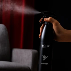 Osoba używa odświeżacza powietrza FOEN Vanilla 500ml, spryskując go z butelki w salonie, co ukazuje zastosowanie produktu do wnętrz mieszkalnych w celu odświeżenia powietrza o zapachu wanilii.